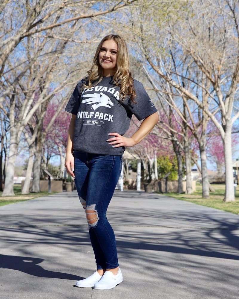 Woman standing on a sidewalk in between to grass areas. She is wearing dark blue ripped jeans and a gray University of Nevada, Reno t-shirt.