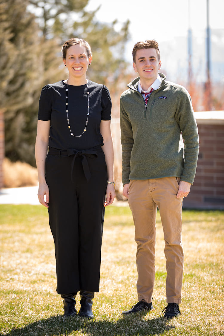 Man and woman standing side by side on part of a grass lawn in front of a partial brown brick wall.