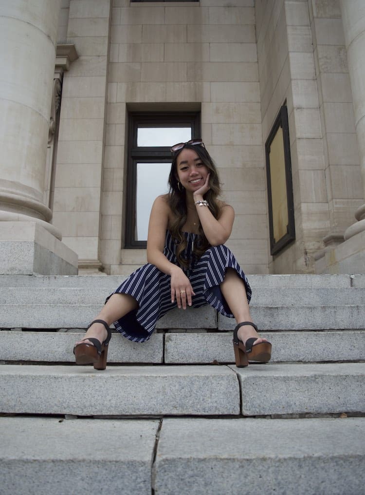 Woman sitting on steps in brown and black heels wearing a blue and white pinstriped strapless jumpsuit that has a bow on the front neckline.