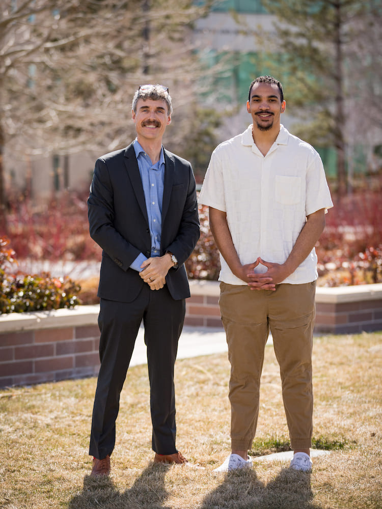 Two men standing on a grass field in front of brown bricked flower beds.