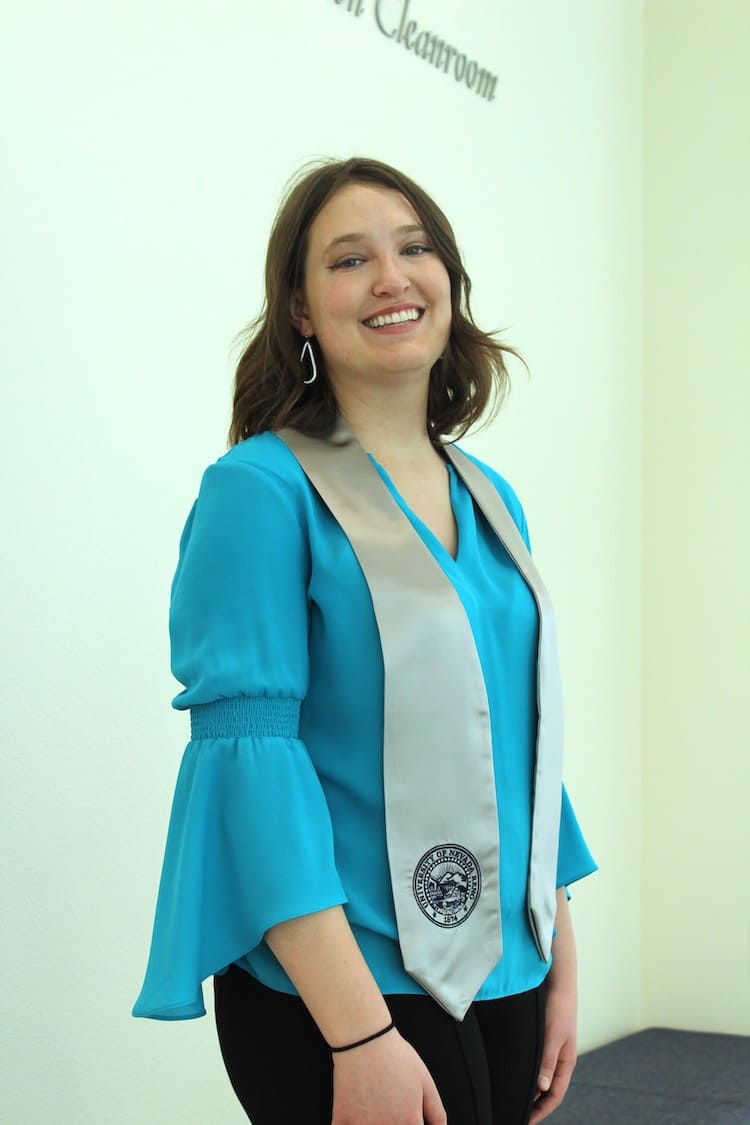 Woman in blue long sleeve shirt posing with a gray graduation stole that features the seal of the University of Nevada, Reno in dark blue.