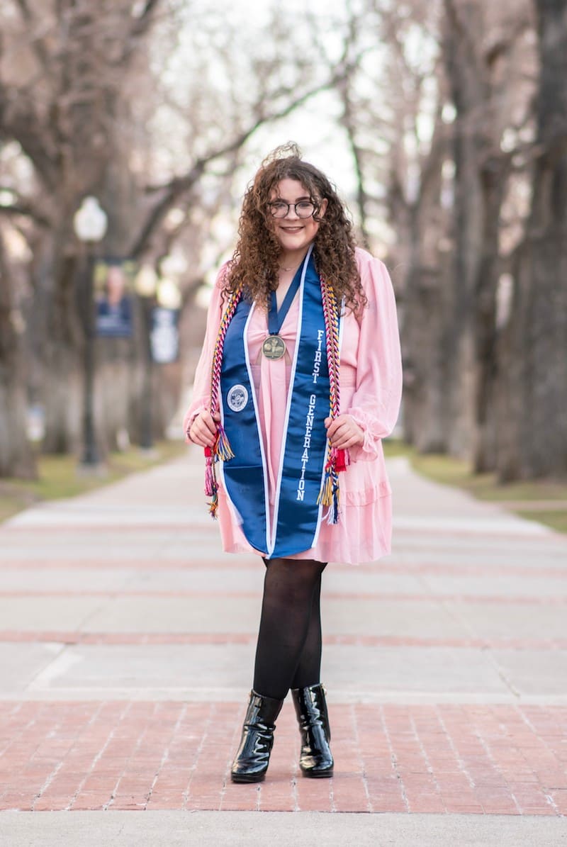 Woman in pink dress with black leather boots and black stockings standing on a bricked sidewalk. She is showcasing her graduation stoles and cords, one of them saying "First Generation."