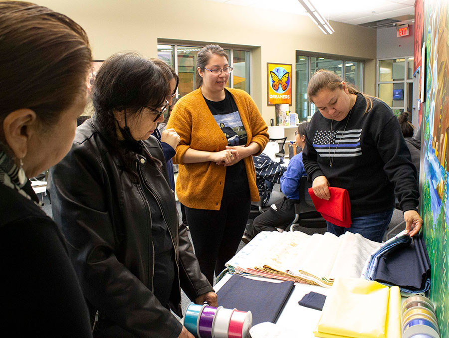 A group of people stand around a table with fabric and ribbon on it. One person to the right digs through various fabric choices and a person to the left lays ribbon on top of fabric to decide on colors.