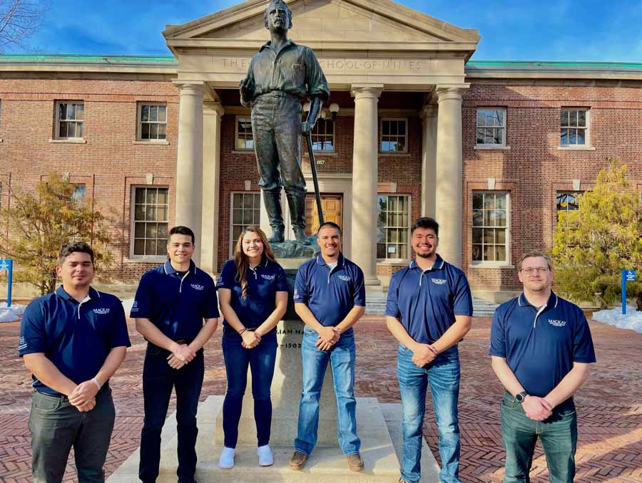 Six people, five of them men, stand in front of the Mackay statue. All are wearing blue Mackay polos.