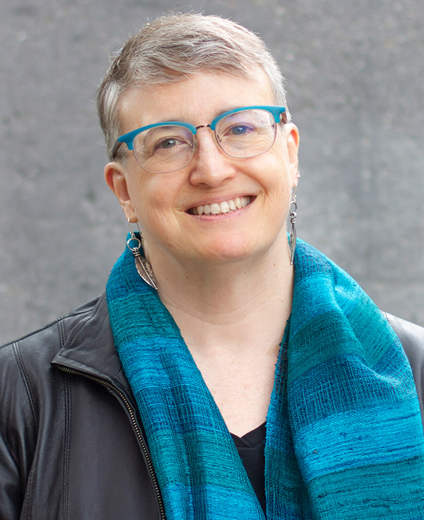 Portrait of Susan Palwick wearing teal rimmed glasses sits for a portrait while wearing a complementary teal scarf and black jacket.