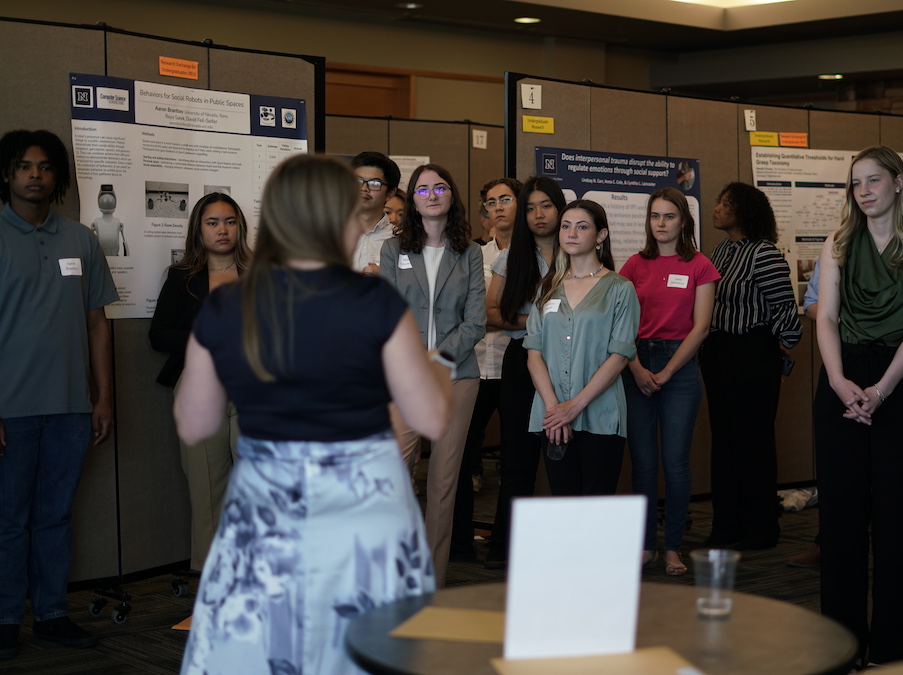 A person's back faces the camera as a group of students politely listen to a presentation while surrounded by posters featuring various research projects.
