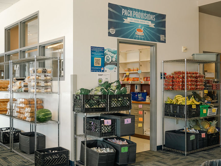 A room with wire shelves to the left of a wall lined with sweets, on the front wall, black crates are filled with vegetables next to a door and a wire shelf filled with strawberries and bananas to the right.