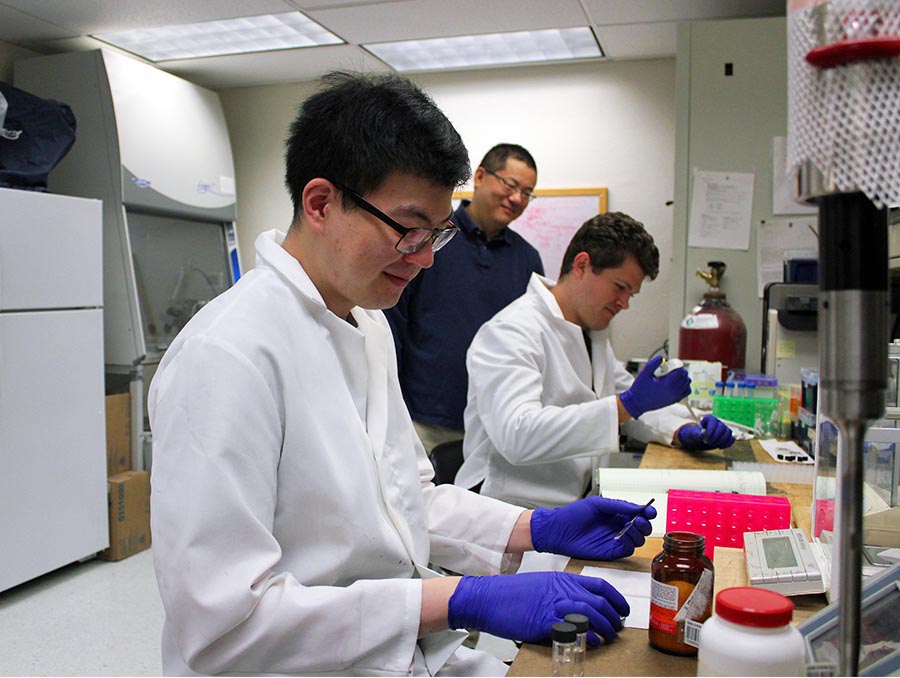 Three people in a lab, two seated and working on something at a table.