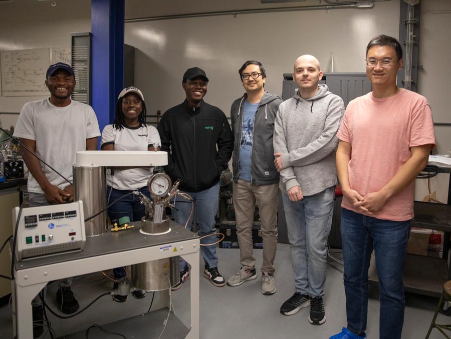 Six people stand in a lab around a piece of equipment with a gauge and control panel on it. They are all smiling.