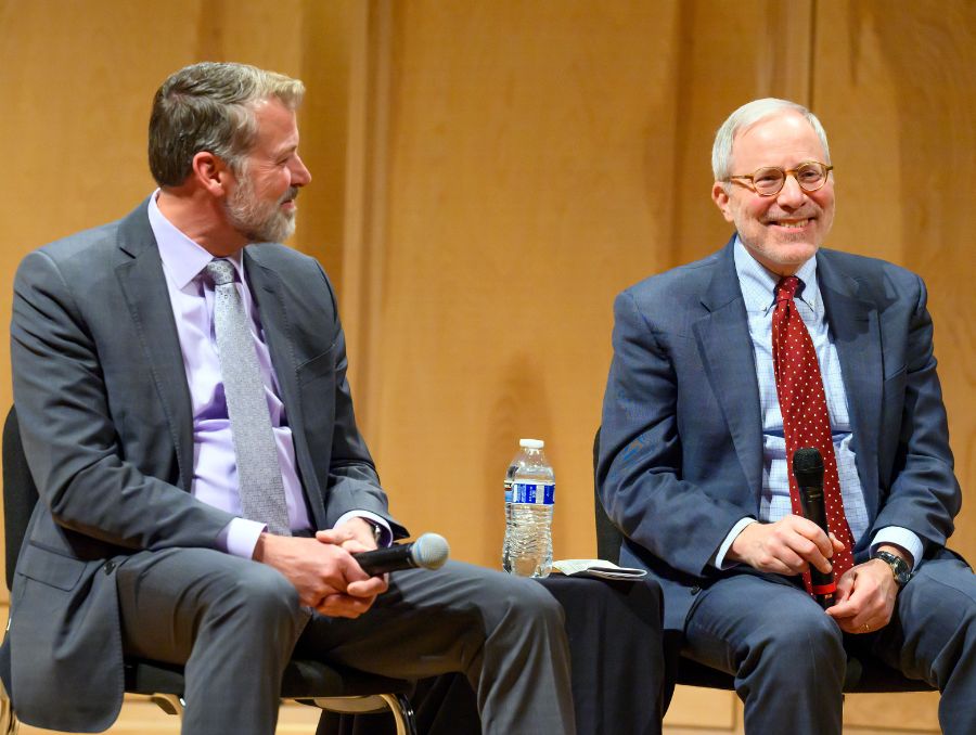 Two men in suits sitting in chairs on stage during panel discussion.