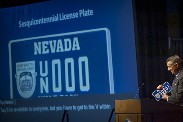 President Sandoval holds a license plate on stage and a close up of the image is projected on screens behind him.