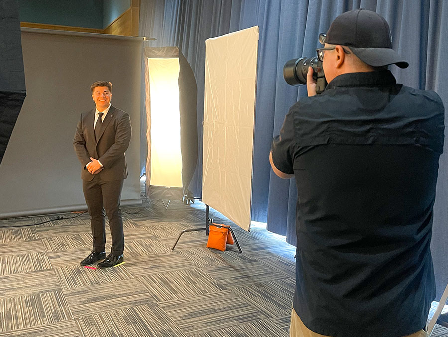 A student poses against a backdrop while a person to the right of the student faces him holding a camera and taking a headshot photo.