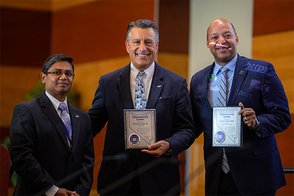 Shamik Sengupta, Brian Sandoval and Erick Jones standing in a row; Sandoval and Jones holding plaques.