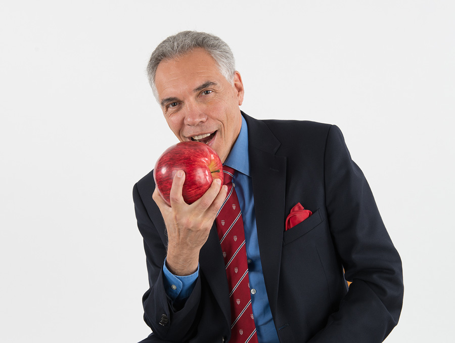 Joe Schwarcz, wearing a suit and tie, holds a large apple and smiles as he pretends to bite into it.