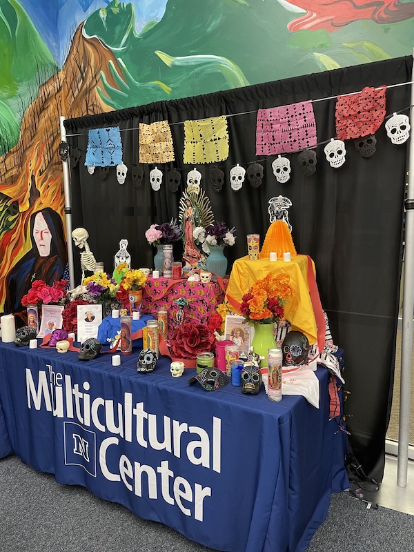 A colorful, traditional altar set up for Dia de Los Muertos with a table cloth that reads Multicultural Center
