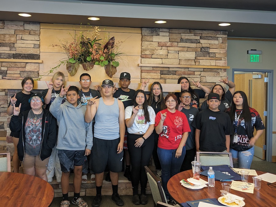 A group of fifteen students poses inside a building, holding up the Wolf Pack hand sign. 