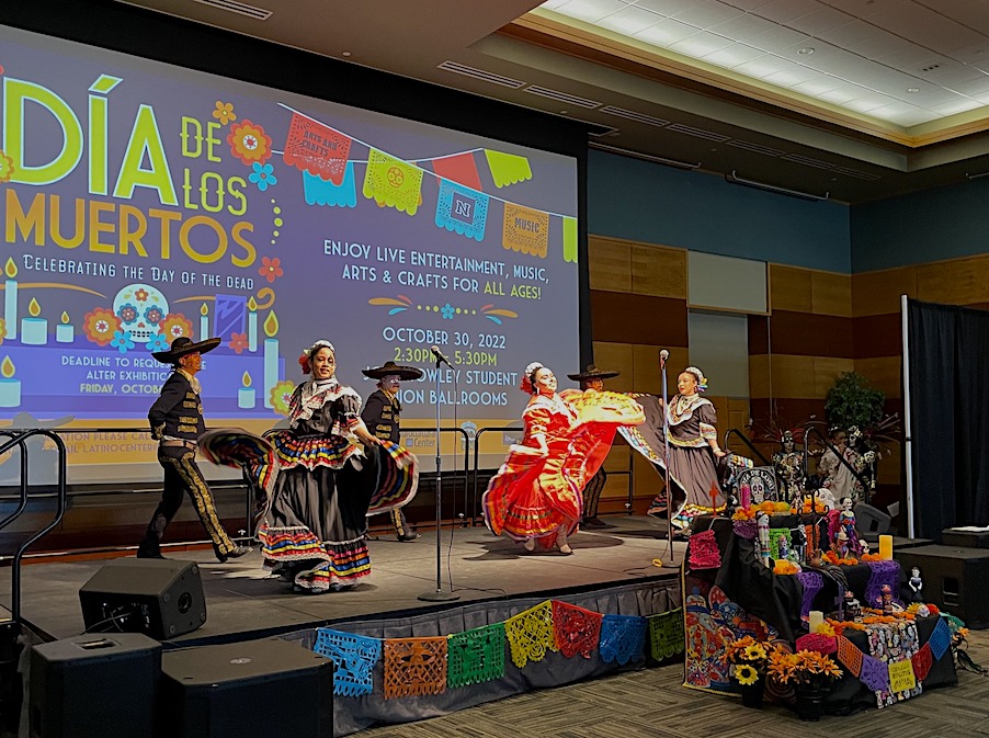 A group of performers on stage as the Dia de los Muertos event in 2022. They wear traditional outfits and dance on stage in front of an altar.