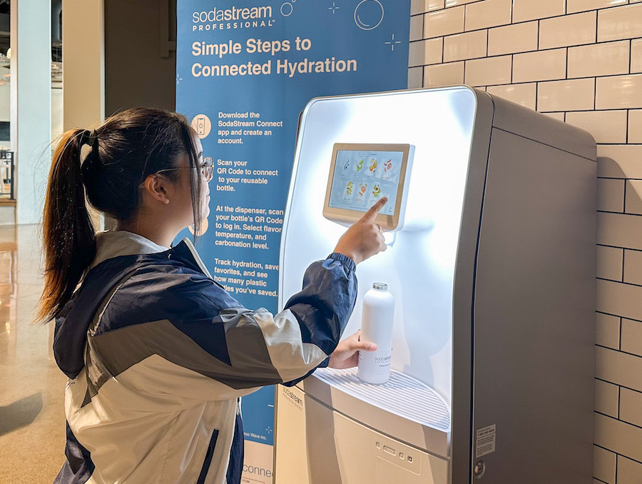 A woman uses a SodaStream machine to make a selection while a banner behind her describes SodaStream saying "Simple Steps to Connected Hydration" and explanatory text below it on how to use the machine, but that text is unfocused.