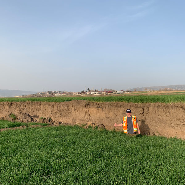 A man in a high-vis vest and a Nevada baseball cap stands in front of a huge piece of uplifted land. A small town is in the background.