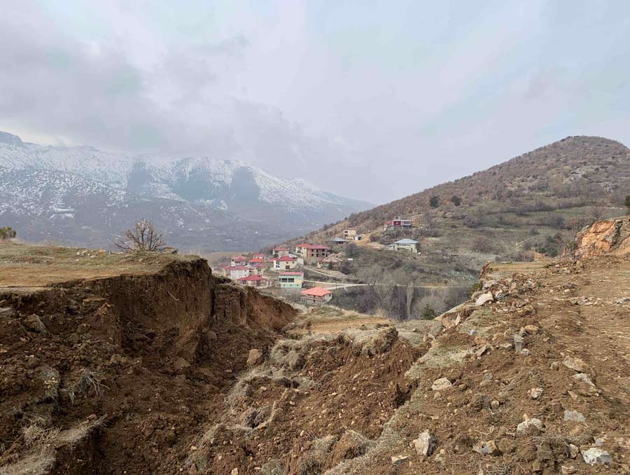 The foreground shows where a block of earth collapsed on a ridge. The background shows a community on a hillside, and in the background are snow-capped mountains.