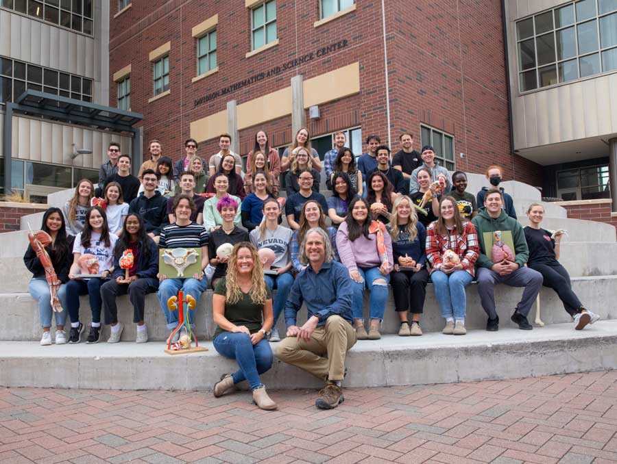 A large group of people sits on the steps outside of the Davidson Mathematics and Science Center. Some of the students are holding biological models.