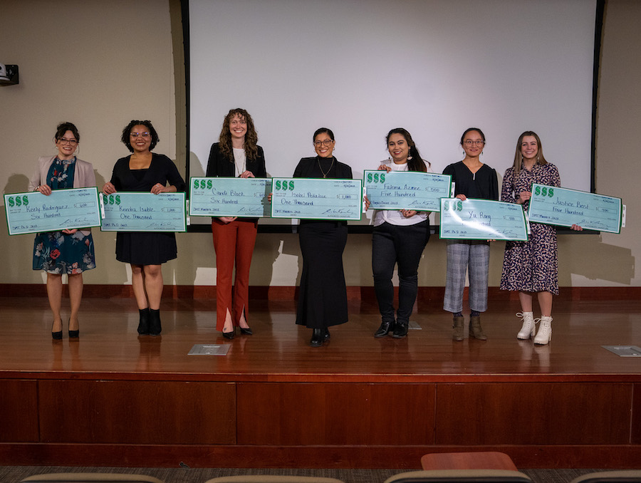 Seven people stand on a stage holding large checks and smiling.