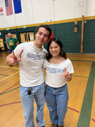 Two college students in the middle school gym wearing University themed t-shirts and holding the wolf pack hand sign.