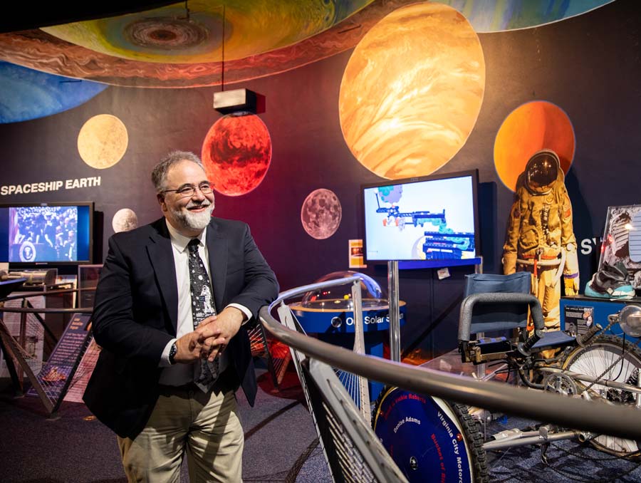 Paul McFarlane leans against a railing in a museum with various artifacts and a mural with the planets painted on the wall behind him.