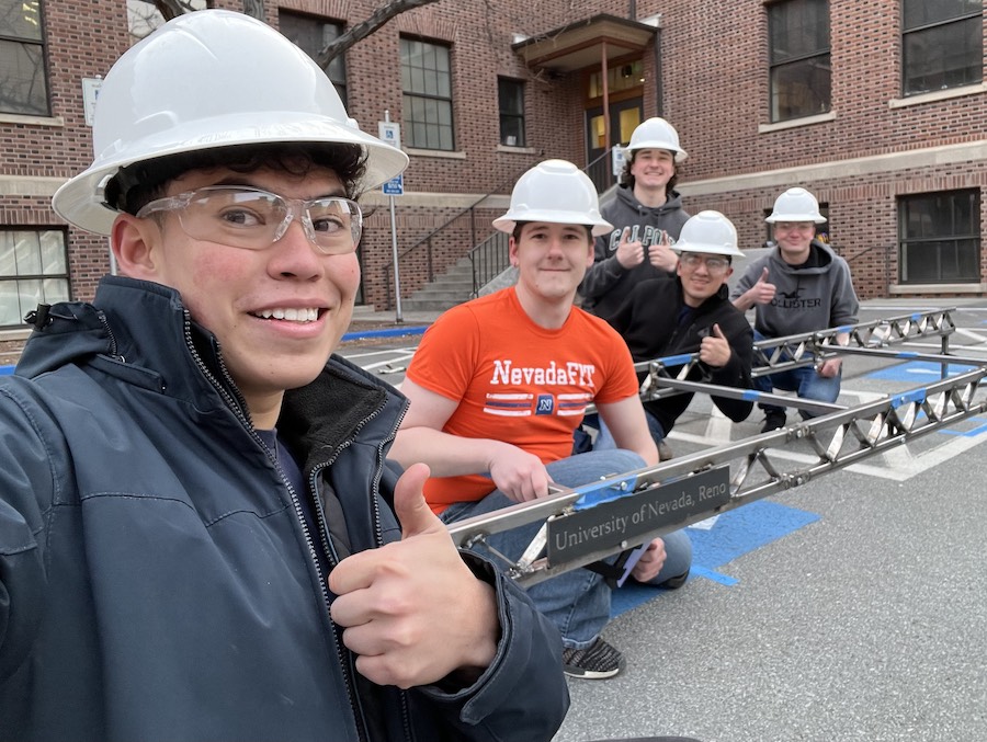 Jose takes a group selfie with four other students, all smiling and wearing hard hats, as they work outside with a metal contraption with a "University of Nevada, Reno" plaque on it. 