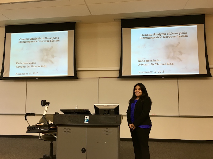 Karla Hernandez stands in front of two PowerPoint screens that display the initial slide of her dissertation research presentation.