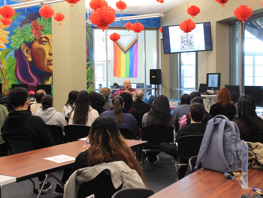 Kalani sits in front of a crowd of students, faculty and staff in the Multicultural Center, speaking into a microphone.