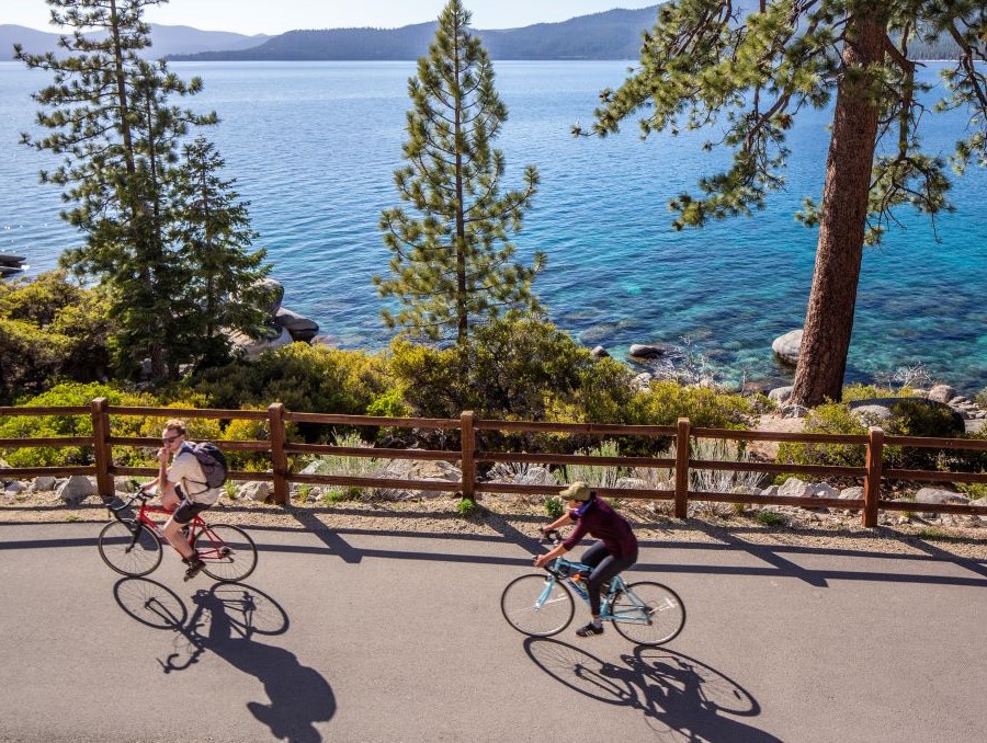 Two bike riders along a paved path with views of pine trees and Lake Tahoe.