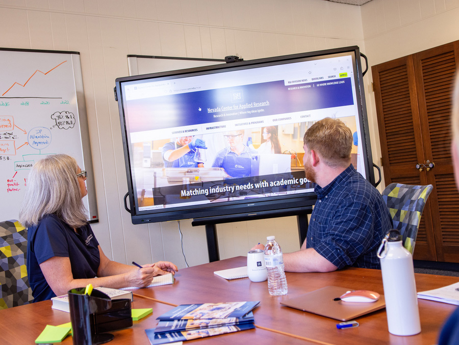 Two people in a meeting, looking at a projector screen with the Nevada Center for Applied Research online page open.