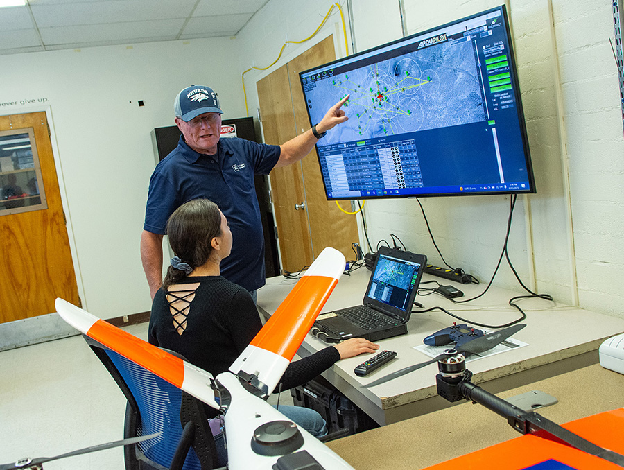UAS Test Site Manager Mark Genung points to a large computer screen explaining Unmanned Aircraft Systems to a person seated.