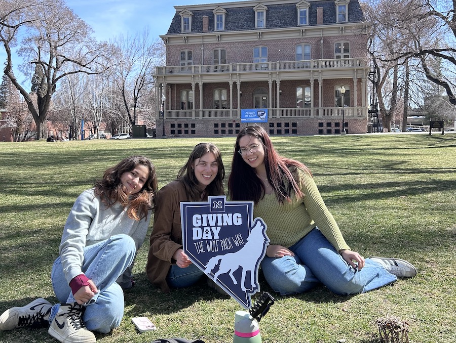 Three students sit in the grass in the quad in front of Morill Hall with a Giving Day The Wolf Pack Way sign.