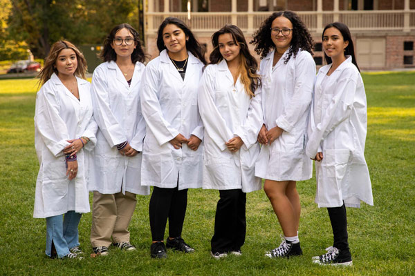 A group of 6 young women wearing lab coats smile on the quad.