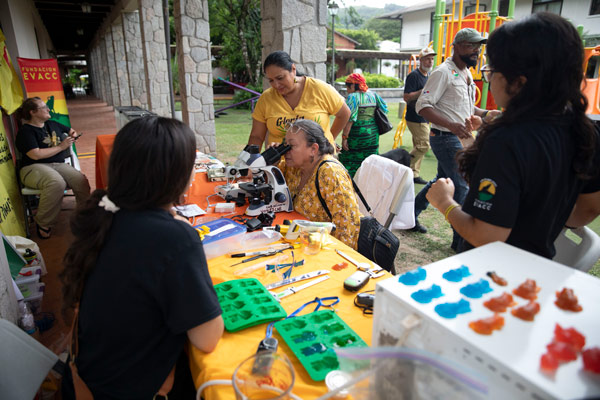 Two women sit down at a microscope on a table with various pieces of scientific equipment laid out as two people in black t-shirts look on.