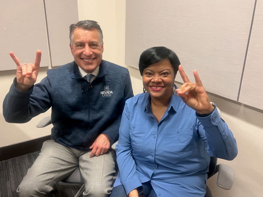 Brian Sandoval sitting next to Melanie Duckworth in the podcasting studio holding up wolf pack hand signs.