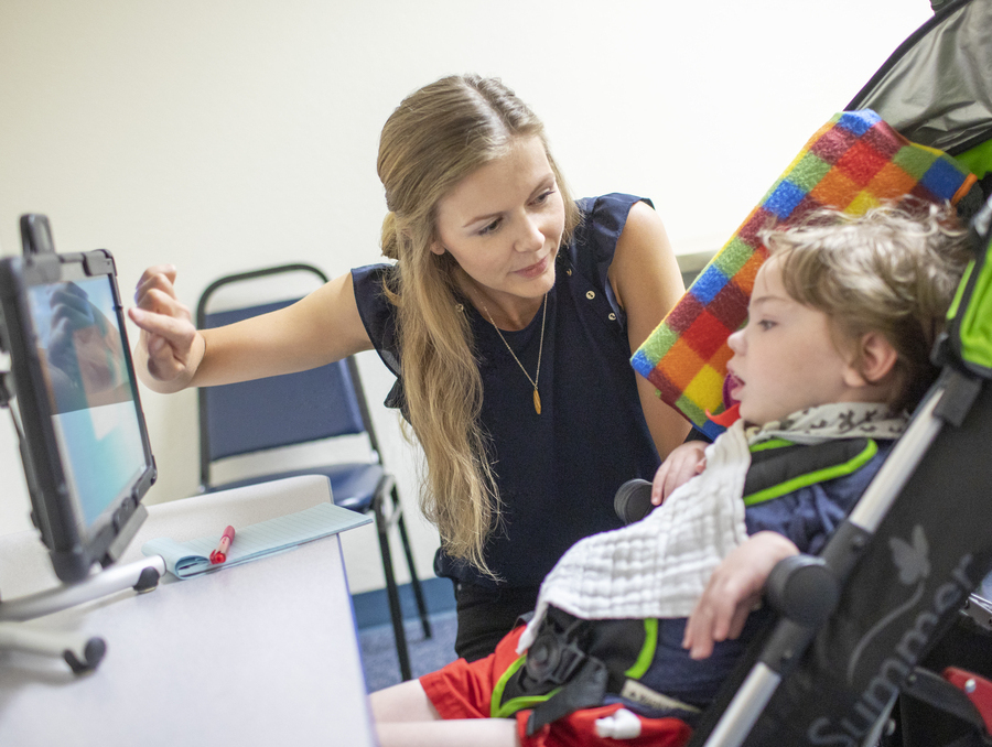 A young woman points to a screen using assistive technology in front of a child in a wheelchair.