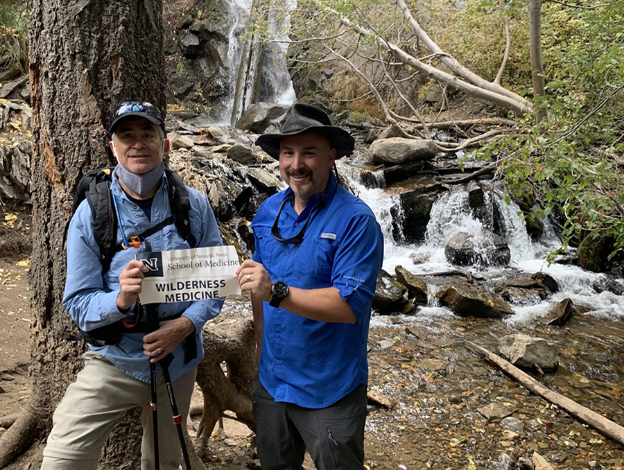David Fiore and Arthur Islas holding up a sign that says "Wilderness Medicine" in a forest next to a waterfall.