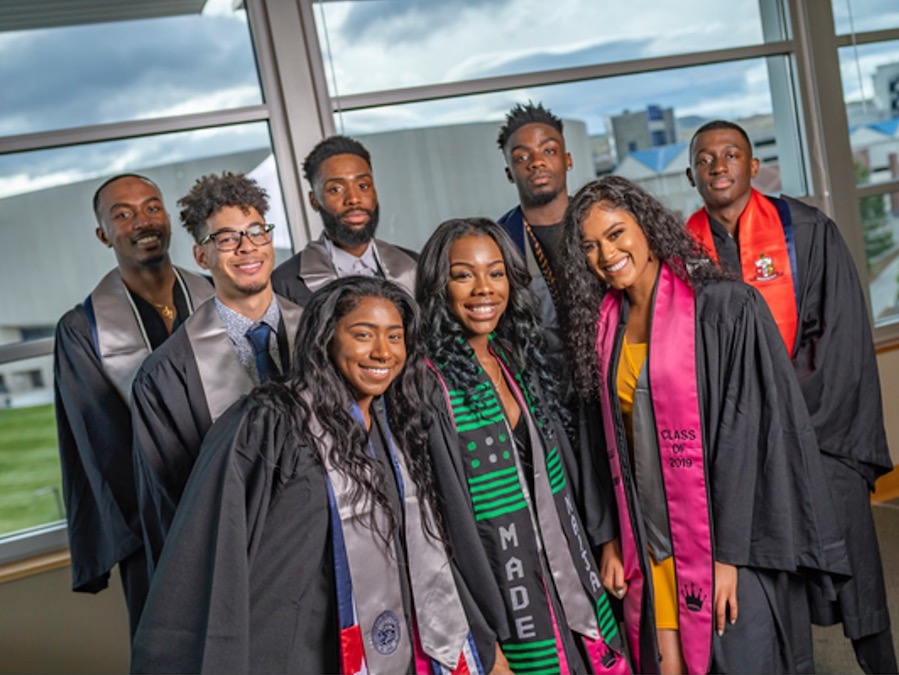 A group of eight students in the African-Diaspora affinity group smiling and posing at the Joe Crowley Student Union with their graduation cap and gowns.