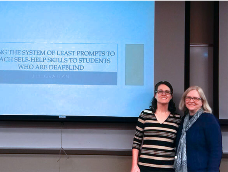 Two people stand near the bottom of a presentation screen and smile.