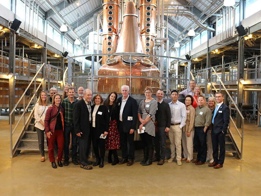 Faculty members pose for a group photo in front of large tanks at a distillery.