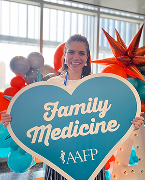Taree smiling and holding a heart-shaped sign that reads "Family Medicine AAFP.”