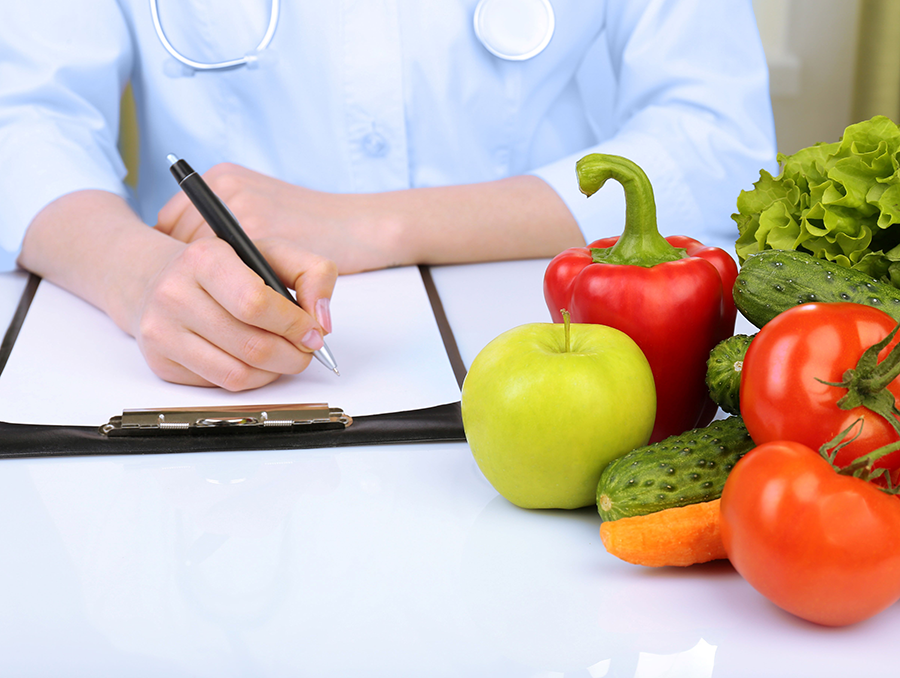 A person sitting down writing with pen with a bunch vegtables on the table.