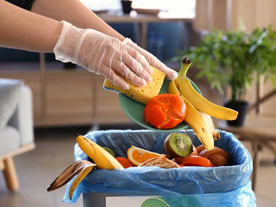 Gloved hands use a kitchen sponge to push food scraps off of a cutting board into the trash. In the pile are scraps of bananas, kiwis, oranges and other fruits.