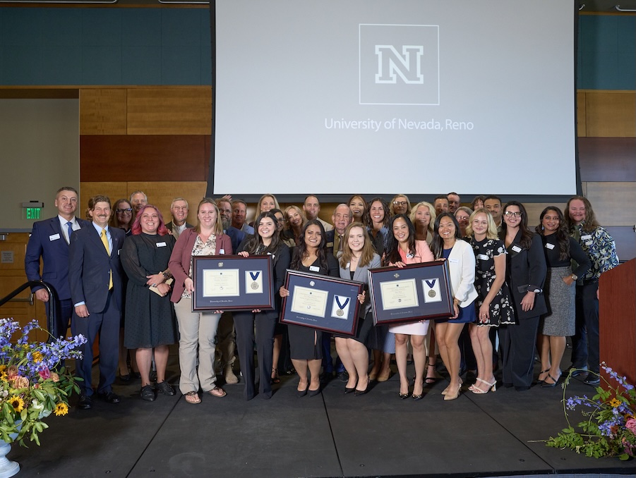 The graduating class in business attire, holding degrees while on stage in a ballroom during the graduation ceremony.