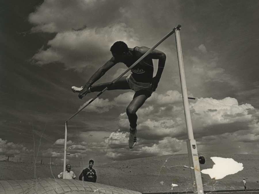 A black and white photo of Otis Burrell wearing a Nevada Track and Field uniform as he jumps over a high bar outside.