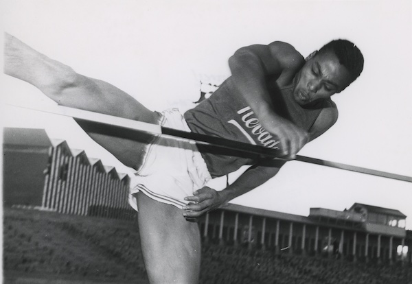 Otis Burrell in a Nevada Track and Field uniform soaring over a high jump pole.