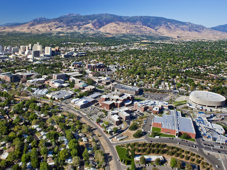 An aerial shot of Reno in the summer with lots of green trees visible and the buildings on the campus of the university as well as those of downtown Reno visible. 